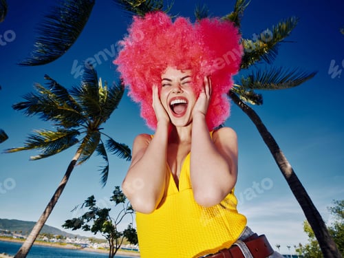 Preview: Pinkhaired woman in sunglasses smiling, standing in front of palm trees with hands on her head