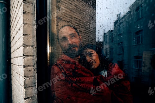 Preview: couple wrapped in a red blanket looking outside from a backyard door window on a grey rainy day