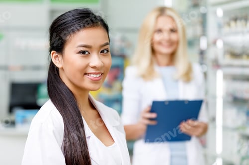 Preview: beautiful female asian pharmacist smiling at camera while colleague with clipboard standing behind