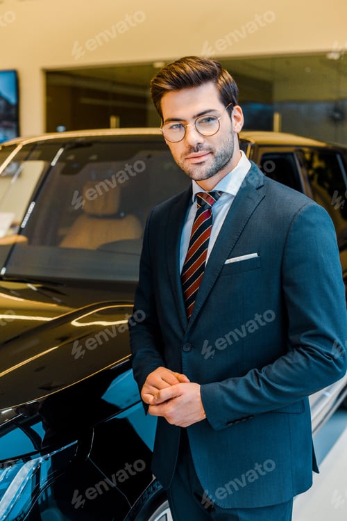 Preview: happy young businessman in eyeglasses posing near car in showroom