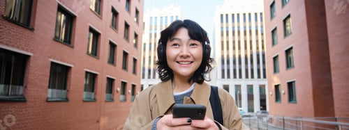 Preview: Portrait of cute girl tourist, korean woman in headphones, looks at mobile phone, uses smartphone