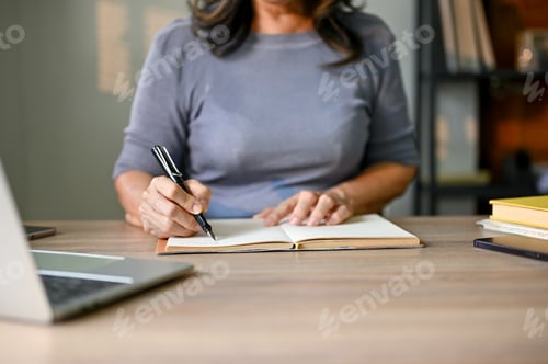 Preview: Cropped image of an Asian-aged businesswoman taking notes on her planner book at her table.