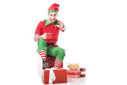 Preview: man in christmas elf costume sitting on pile of presents, holding cup of tea and pointing remote