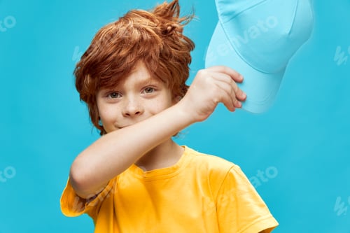 Preview: Red-haired boy with freckles holds a cap in his hands in a yellow t-shirt blue isolated fund