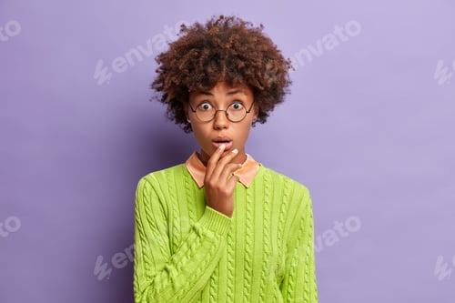 Preview: Excited wondered curly haired woman keeps hand on lips opens mouth from disbelief hears impressive n