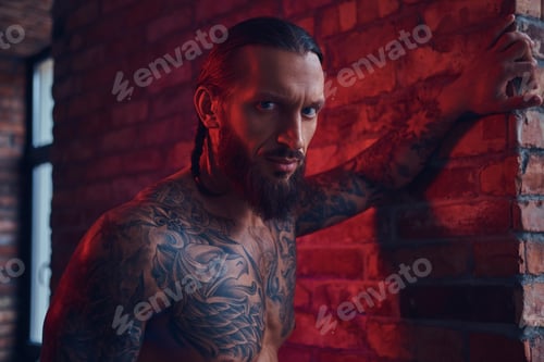 Preview: Handsome tattooed man with a stylish haircut and beard standing against a brick wall