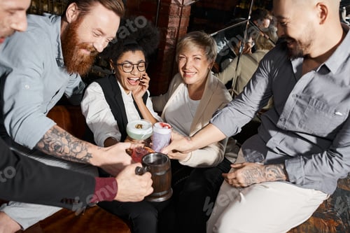Preview: happy women and tattooed men clinking glasses of cocktails in bar, multiethnic friends having fun