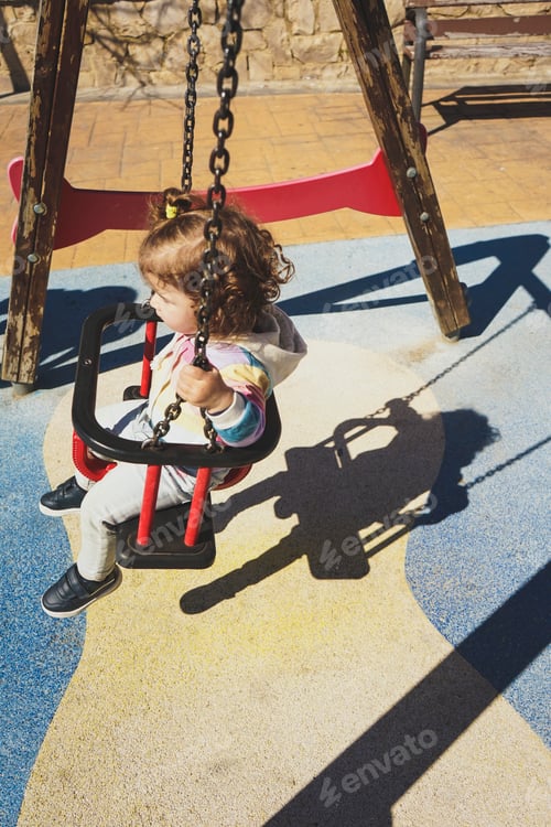 Preview: Artistic and conceptual portrait of a lonely toddler girl alone in a swing ath the playground