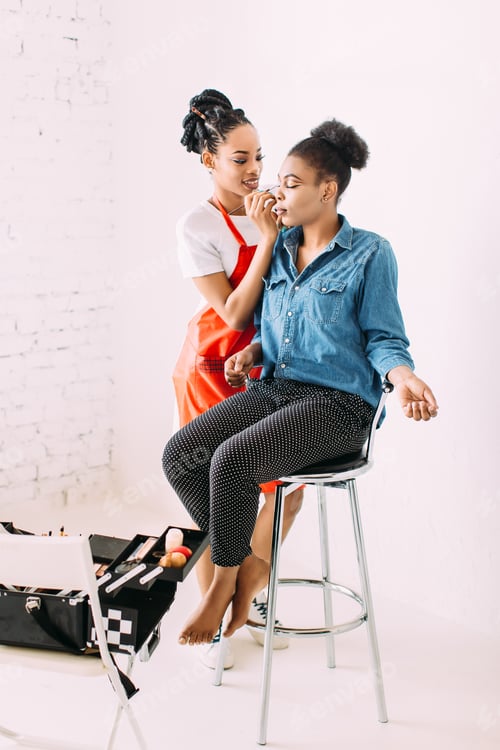 Preview: Young beautiful African American woman applying professional make-up by African make-up artist