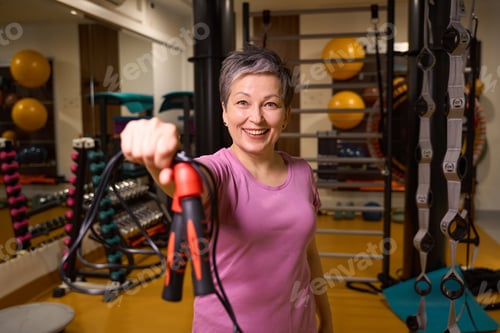 Preview: Mature smiling woman holding skipping rope and looking at camera in gym