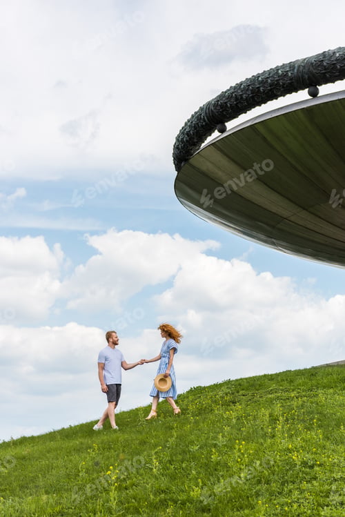 Preview: distant view of redhead couple holding hands on grassy hill near building against blue cloudy sky