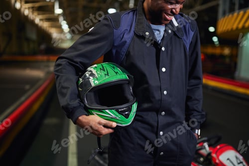 Preview: cheerful african american man in sportswear holding helmet and standing in karting track circuit