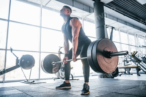 Preview: Strong young man in sportive clothes doing exercises with heavy weights in the gym