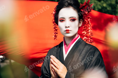 Preview: selective focus of tree branches and beautiful geisha with greeting gesture looking at camera with