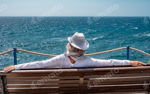 Preview: Back View of Senior Mature Woman with Hat Sitting Outdoor in a Bench Looking at Sea.