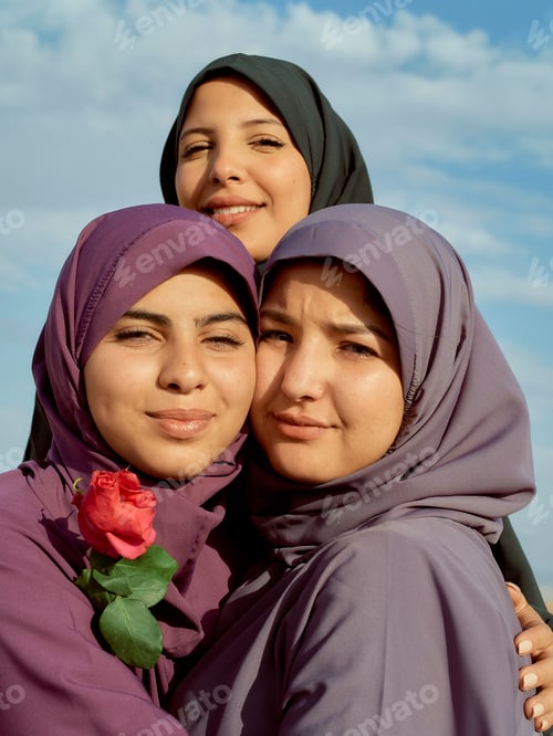 Preview: Three smiling women wearing hijabs embrace against a blue sky, one holding a red rose.