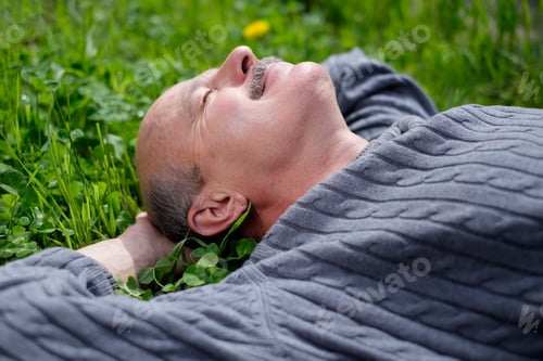 Preview: Mature Happy Man Lying On Green Grass. He holds onion leaf in mouth and closes eyes.