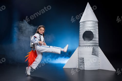 Preview: Happy little girl in astronaut costume jumping near rocket