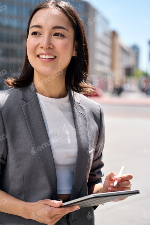 Preview: Young happy Asian business woman standing on city street using digital tablet.