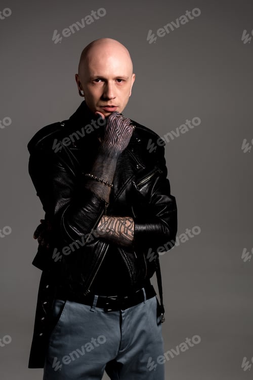 Preview: stylish tattooed man in leather jacket standing with hand on chin and looking at camera isolated on