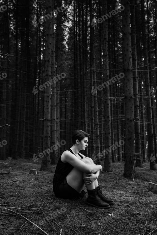 Preview: Grayscale of a young female in a black dress sitting on the ground alone in the forest