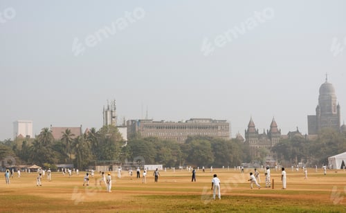 Preview: Cricket players enjoying a game on a hazy morning with historic buildings in the background.