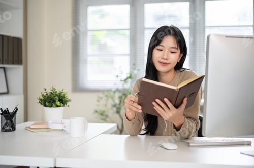 Preview: Pretty asian woman holding pen and looking or reading book aside computer on white table in office.