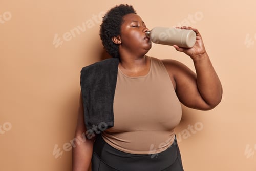 Preview: Sport hydration. Plump dark skinned African woman dressed in tracksuit poses with towel on shoulder