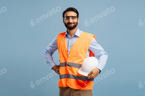 Preview: Smiling, bearded Indian man wearing orange vest and protective eyeglasses holding white hard hat