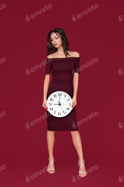 Preview: Pretty young modern business woman in maroon dress holding a clock over red background