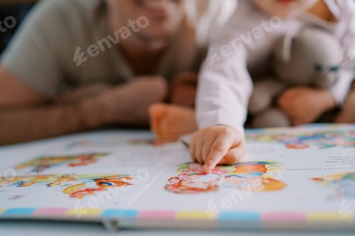 Preview: Little girl points her finger at a colorful picture in a book while sitting with her dad. Cropped