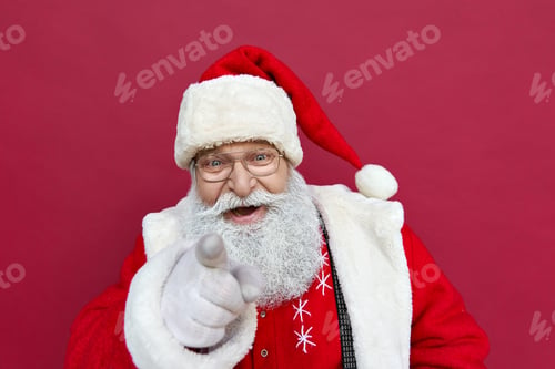 Preview: Happy Santa Claus looking at camera pointing finger at you on red background.