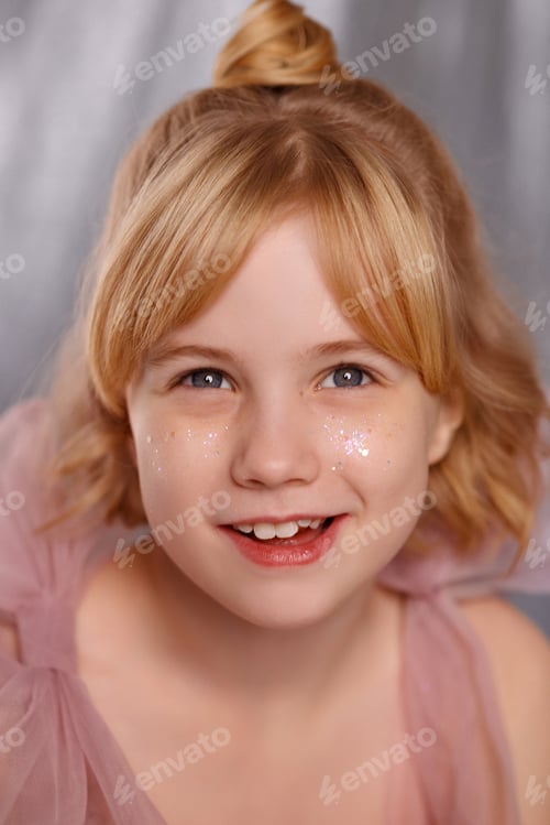 Preview: Smiling blonde girl with glitter on cheeks in pink dress front view on gray background