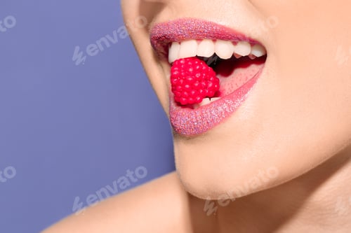 Preview: Beautiful young model with creative makeup holding candy in lips on color background