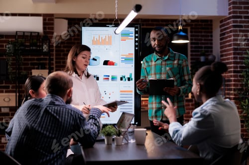 Preview: Two coworkers discussing marketing strategy with colleagues in business office working overtime
