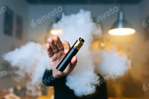 Preview: Young beard man show vaping device on his outstretched hand through a cloud of steam. Selective