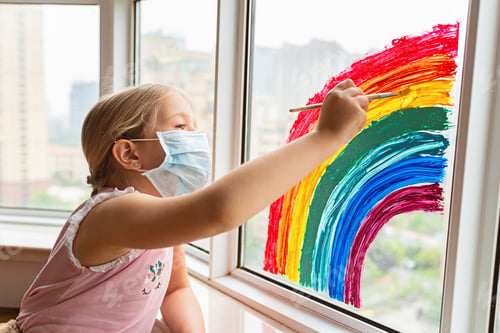 Preview: Little girl in protective facial mask painting rainbow on window during coronavirus quarantine