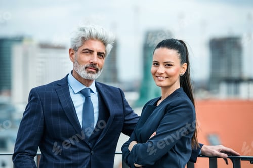 Preview: A businessman and businesswoman standing on a terrace, looking at camera.