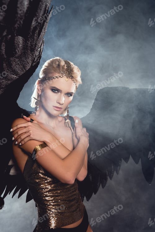 Preview: beautiful young sexy woman with black angel wings and golden accessories posing on dark smoky