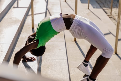 Preview: Young African woman with short hair doing backbend exercise on stairs during outdoor fitness
