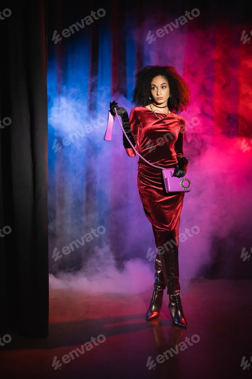 Preview: Trendy african american woman in velvet dress holding telephone on red background with blue light