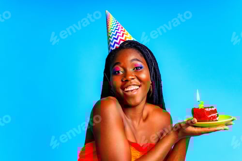 Preview: beautiful afro woman with georgeus makeup wear birthday hat and hold cake in blue studio