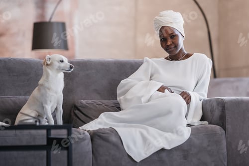 Preview: Confident African young woman in white dress, turban sitting on cozy couch with dog at modern home