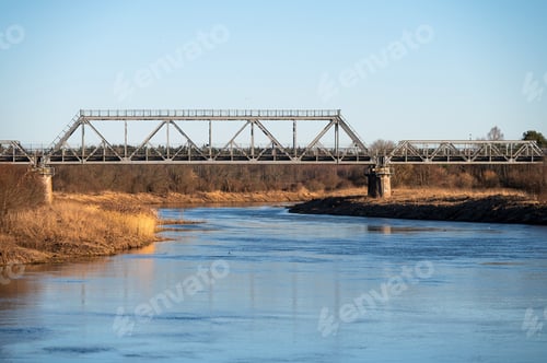 Preview: Metal bridge crosses a calm river surrounded by bare trees and dry grassland in daylight.