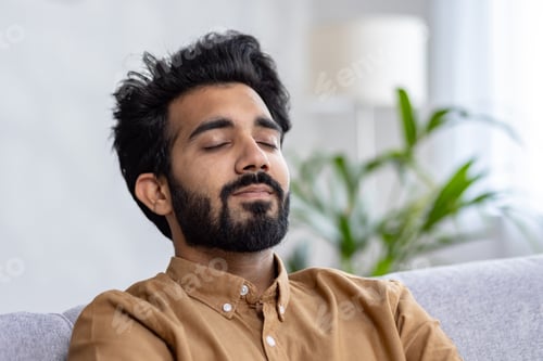 Preview: Portrait of indian bearded man with trendy hairstyle leaning on grey sofa back and closing eyes in