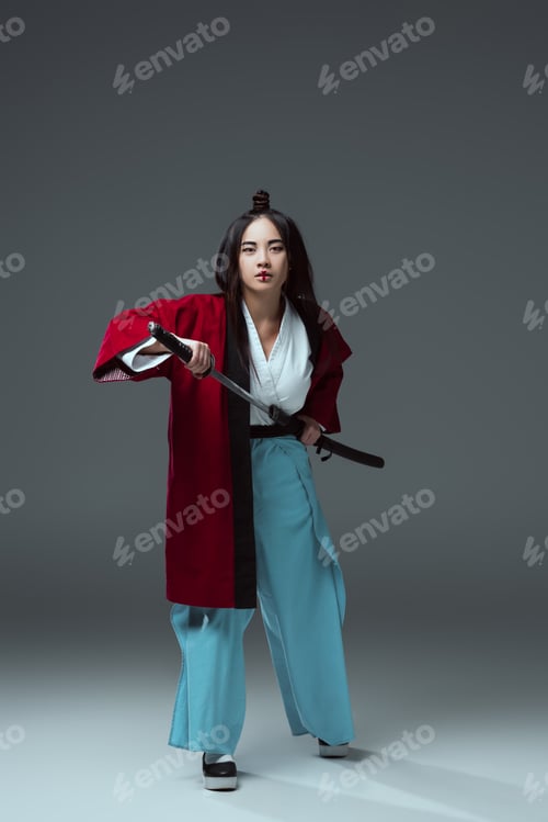 Preview: full length view of young asian woman in kimono holding katana sword and looking at camera on grey