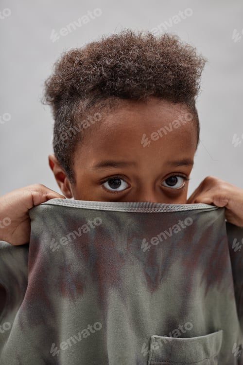 Preview: Little Black Boy Covering Face with T Shirt Collar Peeking out at Camera in Studio