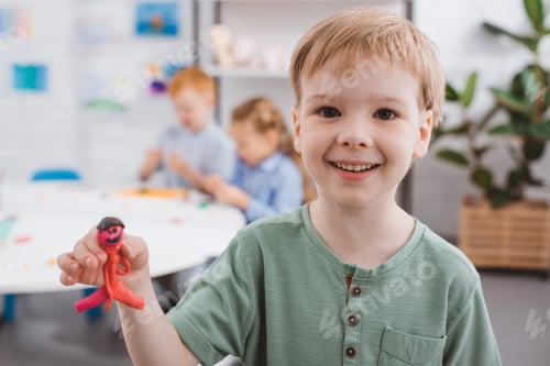 Preview: selective focus of happy kid showing plasticine figure in hand with classmates behind in classroom