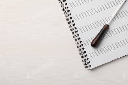 Preview: Conductor's baton and sheet music book on white wooden table, top view. Space for text
