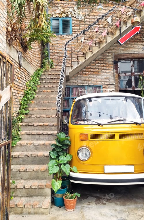 Preview: Yellow vintage van parked next to a brick building with stairs and green plants.
